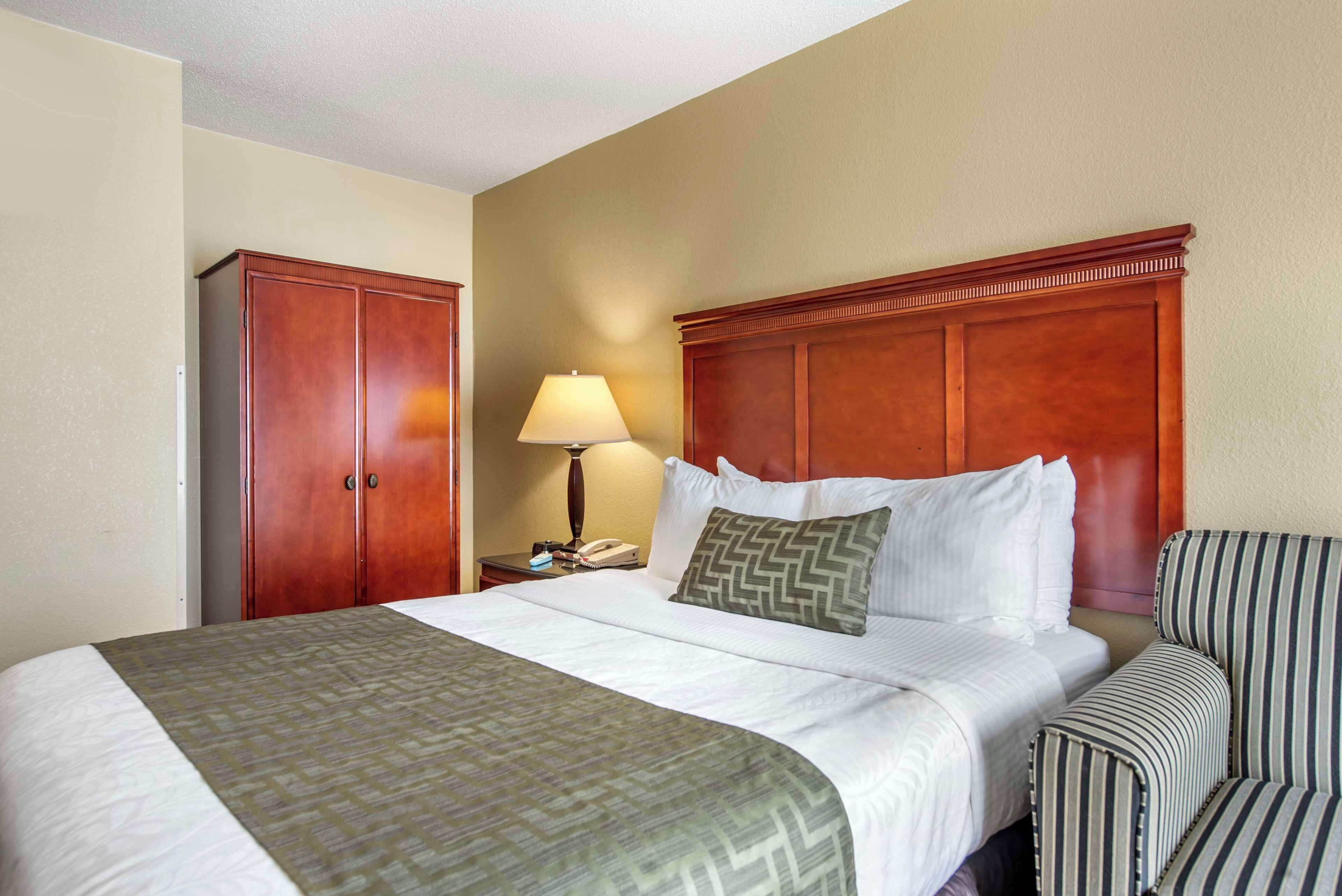 A hotel room with a large bed, a wooden headboard, a bedside table with a lamp, and a striped armchair.