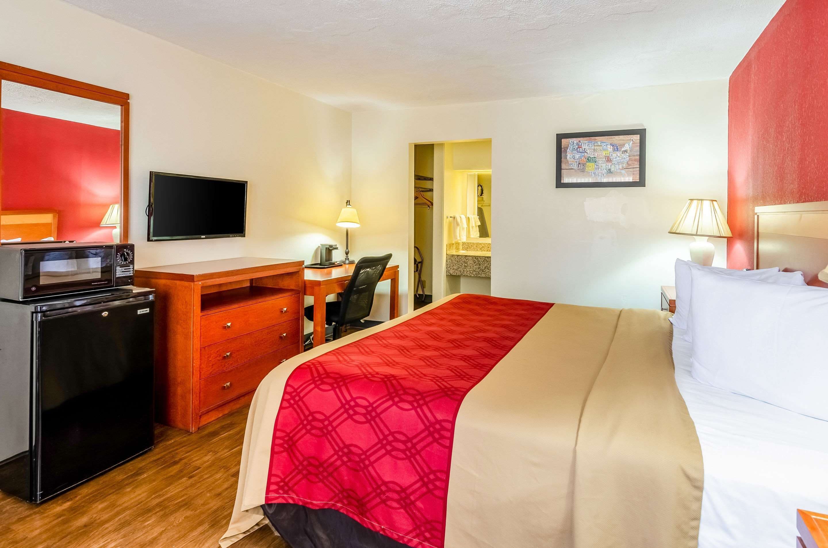 A hotel room with a bed, a desk with a chair, a television, a refrigerator, and a bathroom visible through an open door.