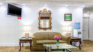 Eine Lobby mit einem braunen, gemusterten Sofa, einem Glastisch mit Blumenvase, zwei Nachttischen mit Lampen, einem großen, verzierten Spiegel, einem Flachbildfernseher und einem Willkommensschild.