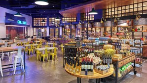 A café with tables and chairs, a counter with various items, and a display of bottles.