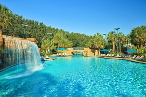 A large outdoor swimming pool with a waterfall feature, surrounded by palm trees and lounge chairs.