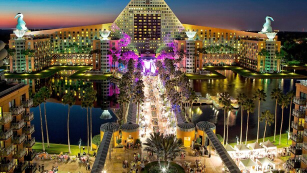 A large hotel with a distinctive pyramid-shaped building, a central courtyard with palm trees, and a body of water reflecting the structure.