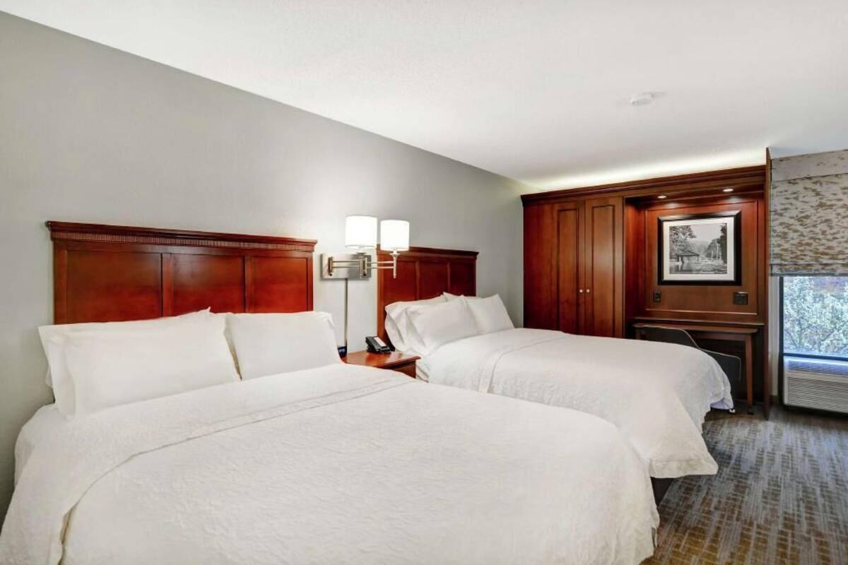 A hotel room with two beds, a wooden headboard, a bedside table with a lamp, and a window with a view.