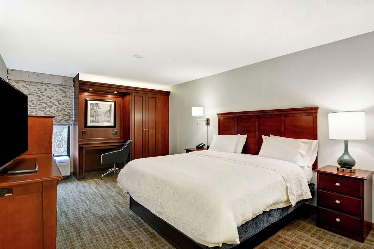 A hotel room with a large bed, a desk, a chair, a TV, and a lamp.