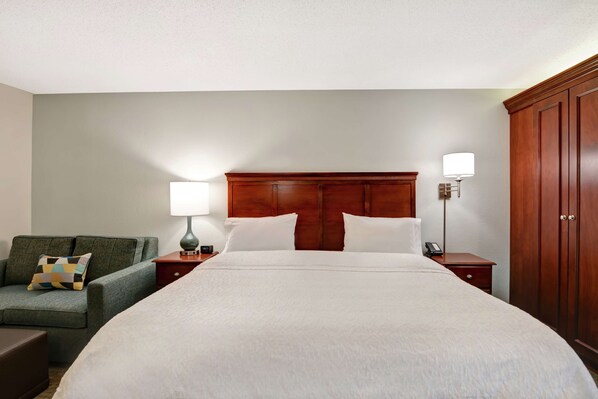 Quarto de hotel com uma cama grande, cabeceira de madeira, sofá verde com almofada estampada, duas mesas de cabeceira com luminárias e um guarda-roupa.