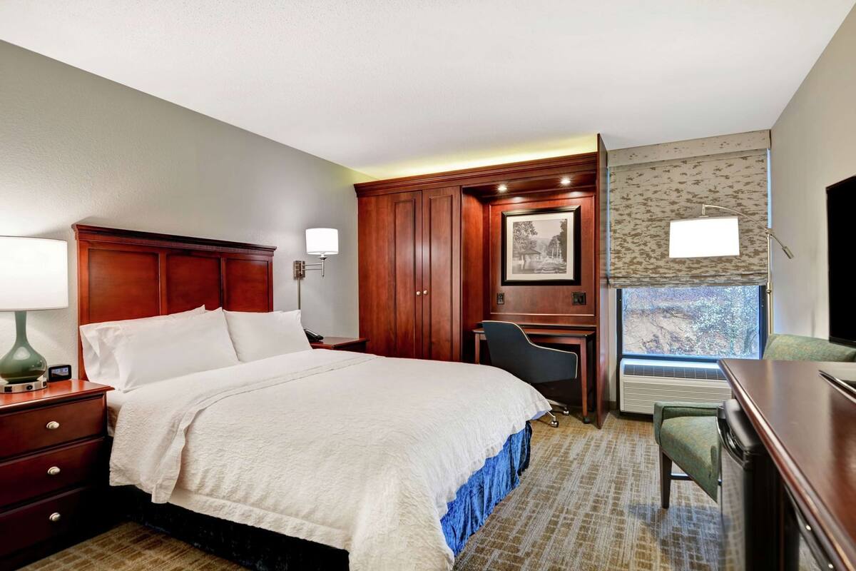 A hotel room with a bed, desk, chair, lamp, and a window with a view.