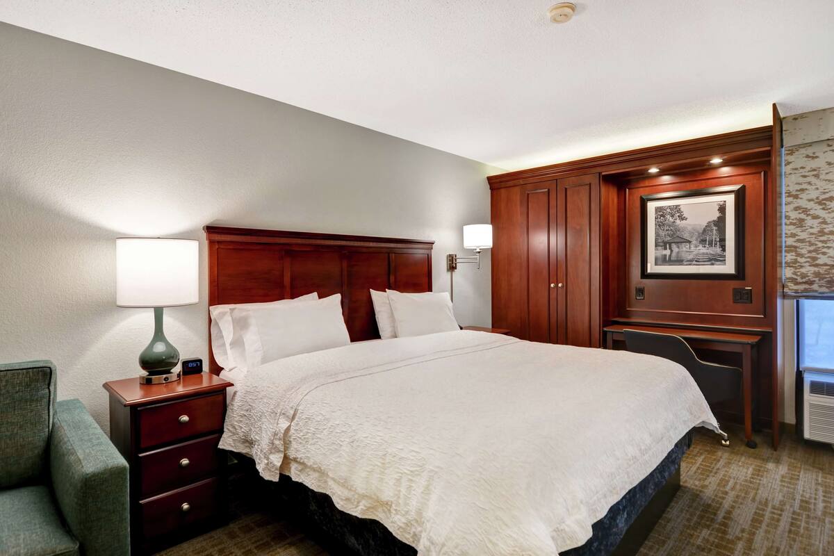 A hotel room with a large bed, a nightstand with a lamp, a desk with a chair, and a framed picture on the wall.
