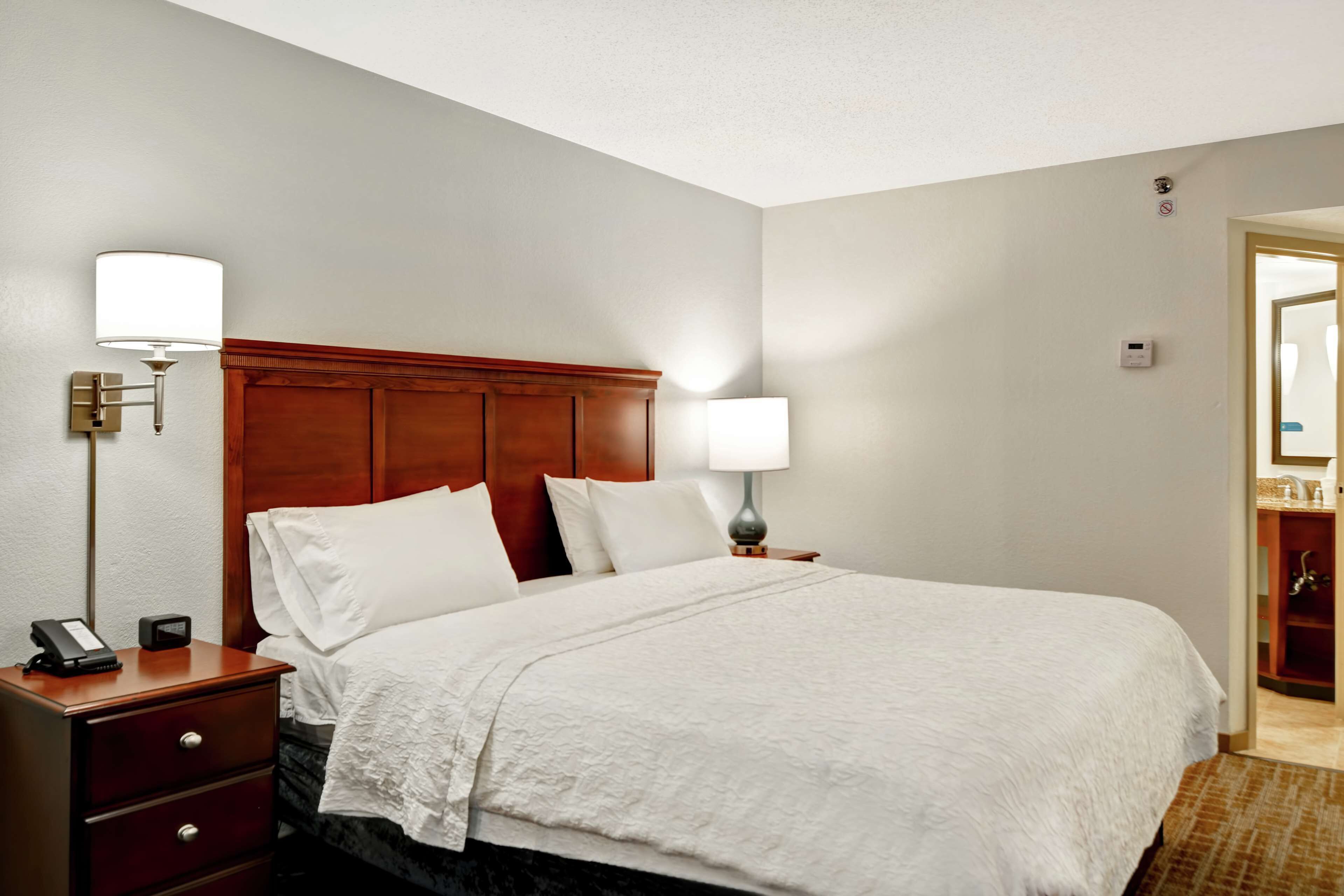 Habitación de hotel con cabecera de madera, cama con ropa de cama blanca, lámpara de noche, mesita de noche con teléfono y puerta que da a un baño.