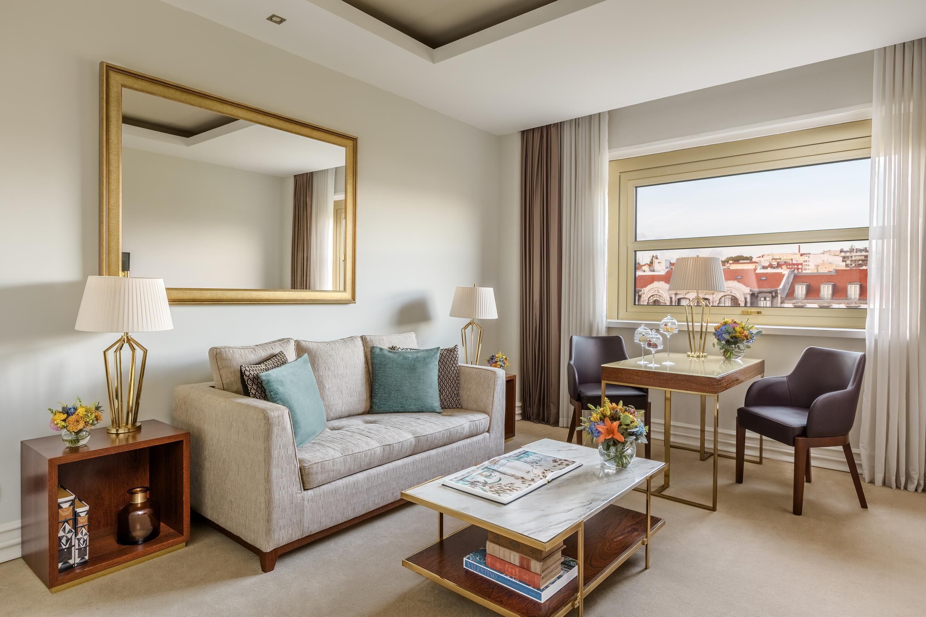 Habitación de hotel con sofá, una mesa de centro, una mesa de comedor con sillas, un espejo grande y un ventanal con vista a la ciudad.