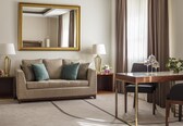 A hotel room with a sofa, two lamps, a dining table with chairs, a mirror, and curtains.