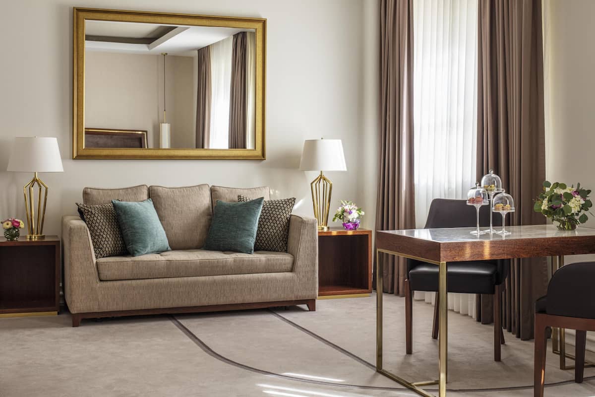 Ein Hotelzimmer mit Sofa, zwei Lampen, einem Esstisch mit Stühlen, einem Spiegel und Vorhängen.