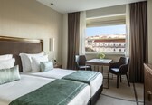 A hotel room with two beds, a small table, two chairs, a desk, and a view of buildings.