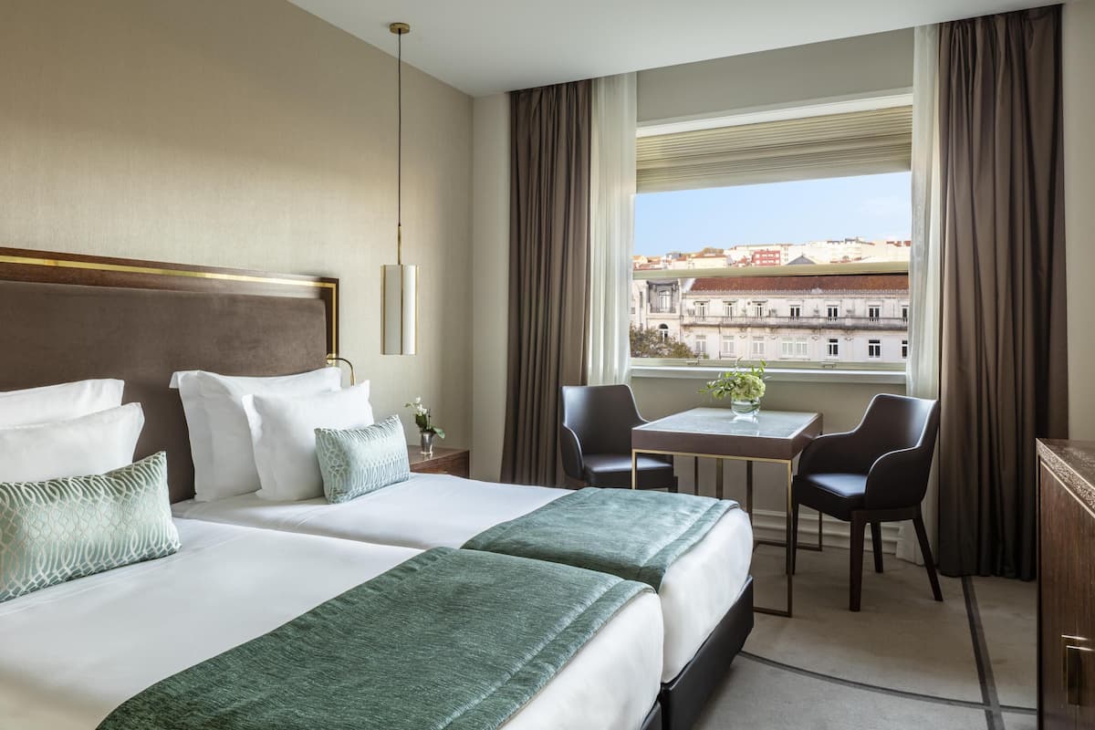 Ein Hotelzimmer mit zwei Betten, einem kleinen Tisch, zwei Stühlen, einem Schreibtisch und Blick auf Gebäude.