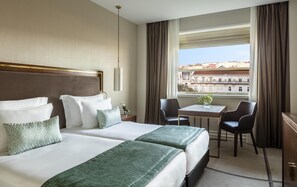 A hotel room with two beds, a small table, two chairs, a desk, and a view of buildings.