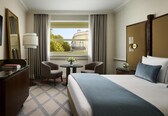 A hotel room with a bed, two chairs, a small table with a vase, a desk with a lamp, and a window with curtains.