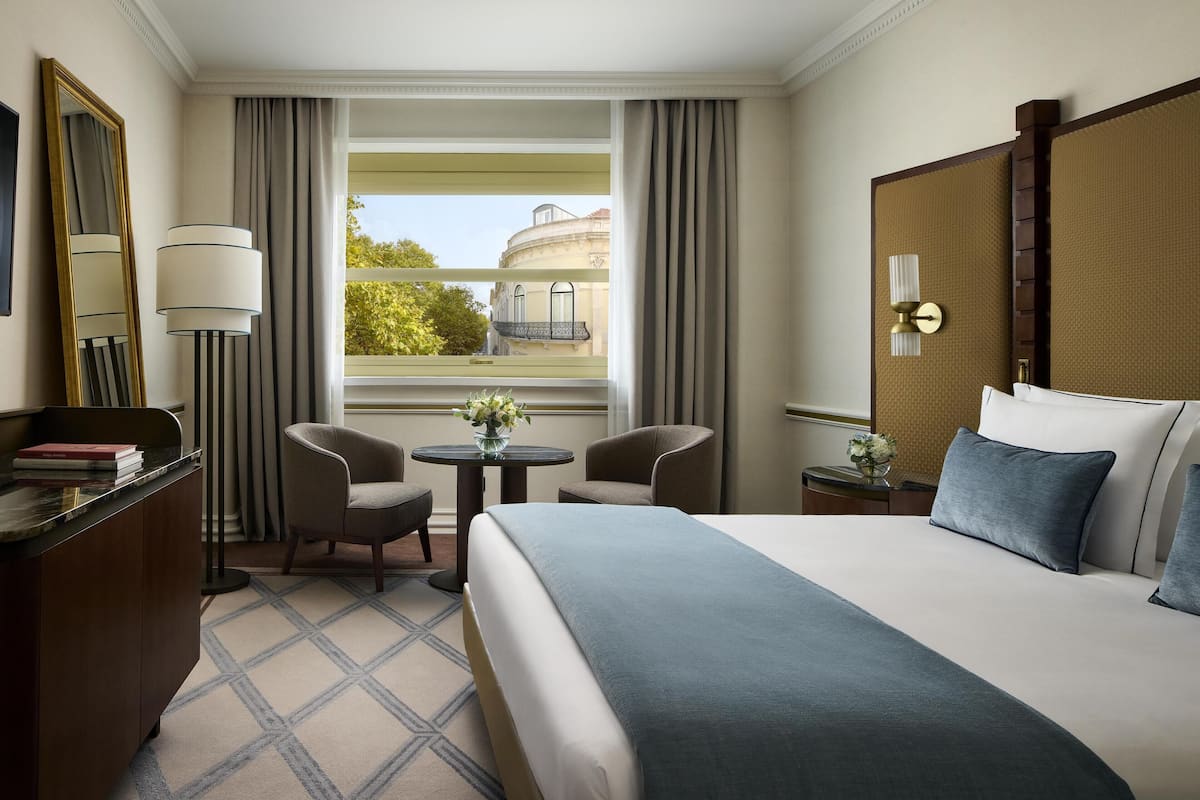 Ein Hotelzimmer mit einem Bett, zwei Stühlen, einem kleinen Tisch mit einer Vase, einem Schreibtisch mit einer Lampe und einem Fenster mit Vorhängen.