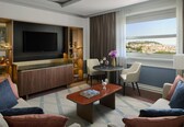 A modern hotel room with a TV, seating area, dining table, and a view of a cityscape.