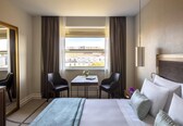 A hotel room with a bed, two chairs, a small table with a vase, and a window with curtains.