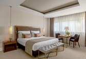 A hotel room with a large bed, bedside lamps, a desk with a chair, a bench, and a window with curtains.