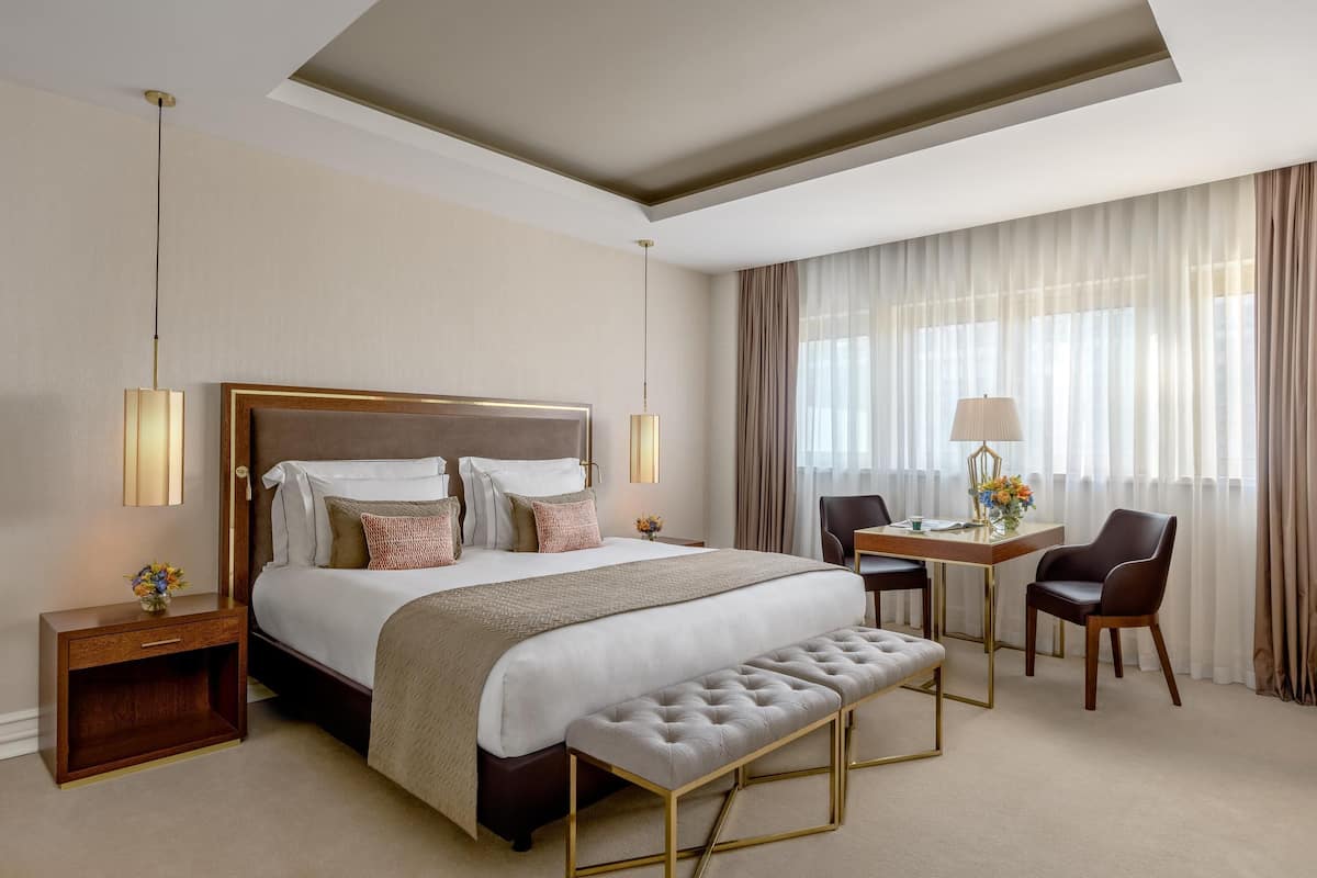 Ein Hotelzimmer mit einem großen Bett, Nachttischlampen, einem Schreibtisch mit Stuhl, einer Bank und einem Fenster mit Vorhängen.