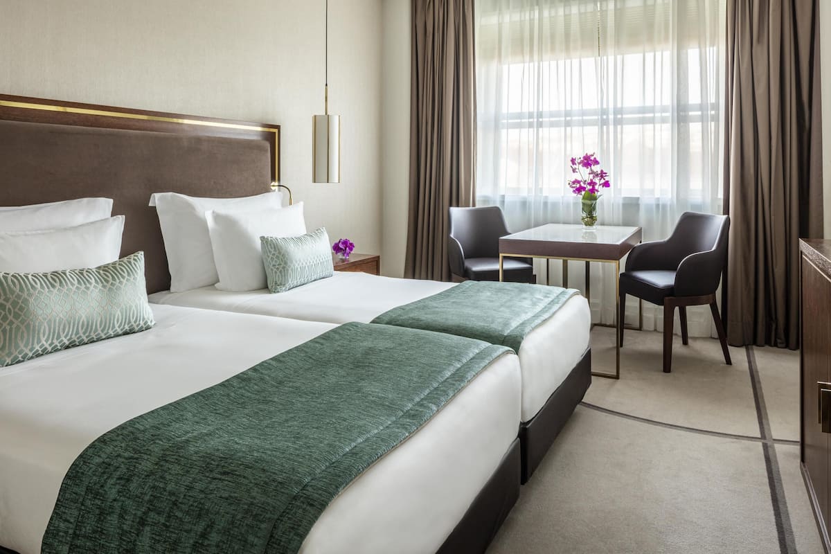 A hotel room with two beds, a desk, chairs, a vase with flowers, and a window with curtains.