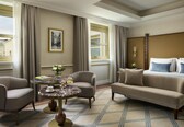 A hotel room with a bed, a sofa, two armchairs, a round table with a vase of flowers, and a framed picture on the wall.