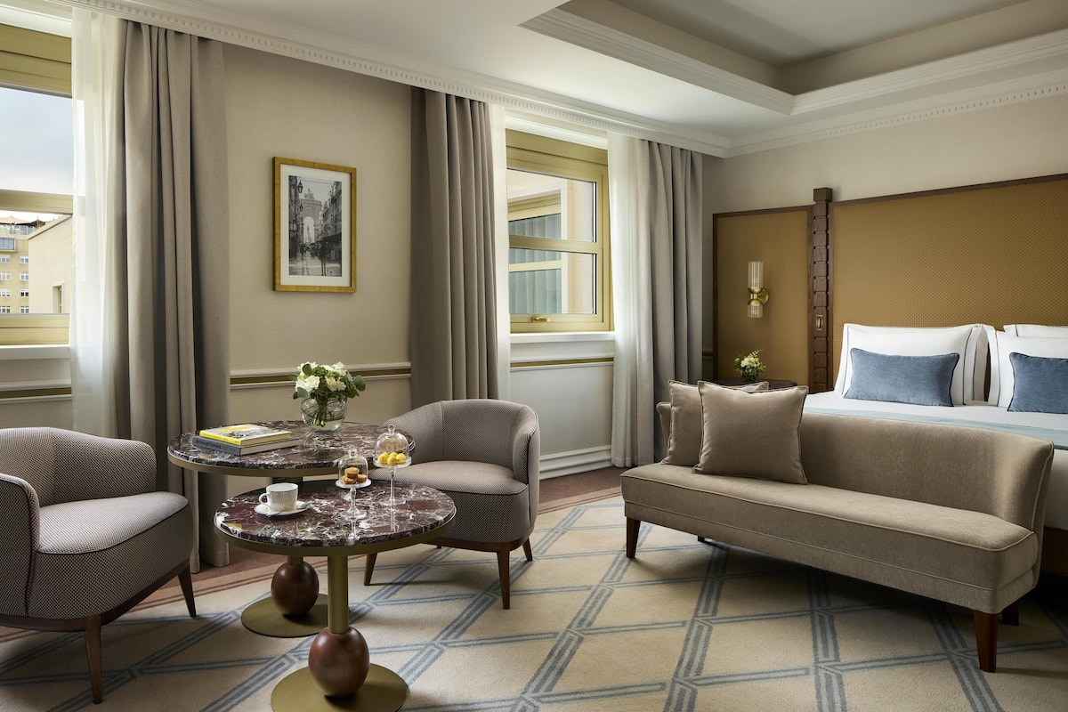 Ein Hotelzimmer mit einem Bett, einem Schlafsofa, zwei Sesseln, einem runden Tisch mit Blumen und einem gerahmten Bild an der Wand.