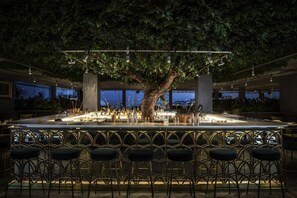 A bar area with a large tree, multiple bar stools, and a view of the cityscape through large windows. - Tivoli Avenida Liberdade Lisboa – A Leading hotel of the world (Lisbon)