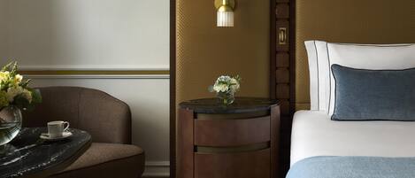 Habitación de hotel con cama, una silla, una mesita con un florero y una lámpara de pared.
