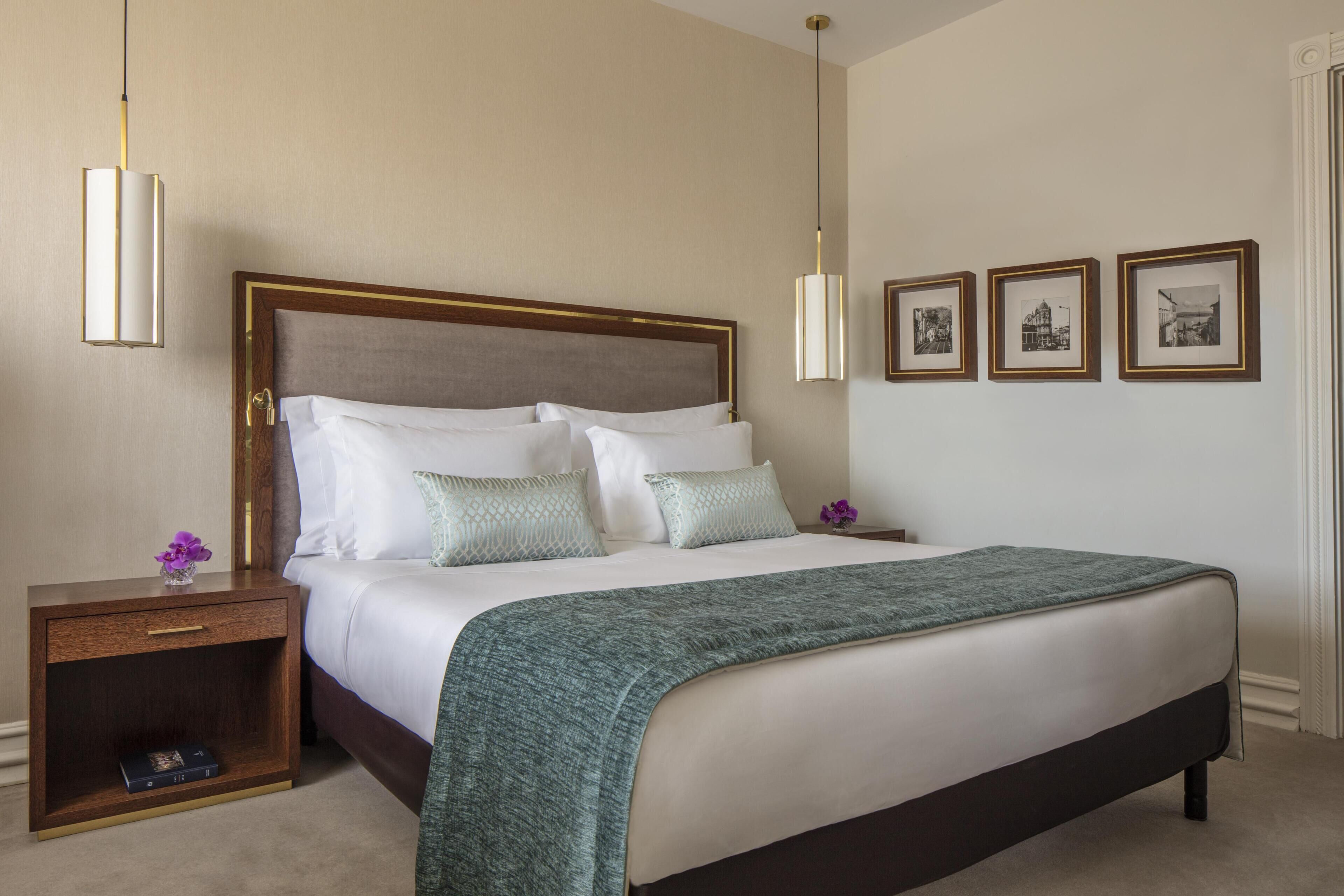 Una cama bien hecha con cabecera de madera, una mesita de noche con cajón y una pared con tres cuadros enmarcados.