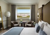 A hotel room with a bed, two armchairs, a small table with a vase, a lamp, and a view of buildings through a window.