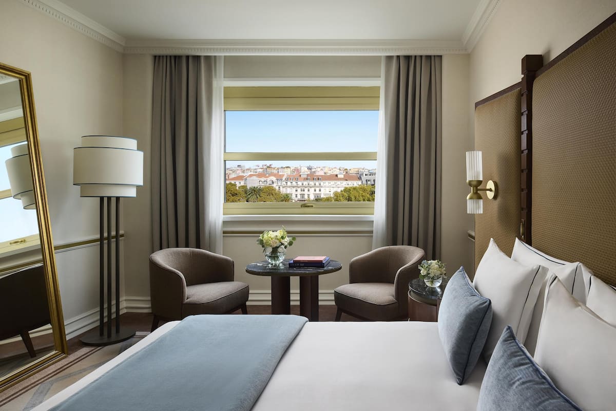 Ein Hotelzimmer mit einem Bett, zwei Sesseln, einem kleinen Tisch mit einer Vase, einer Lampe und Blick auf Gebäude durch ein Fenster.