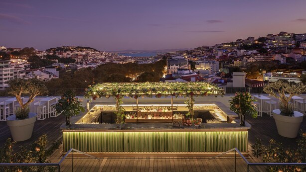Un bar en la azotea con mesa de billar, zona de bar y asientos, con vistas a un paisaje urbano al atardecer.