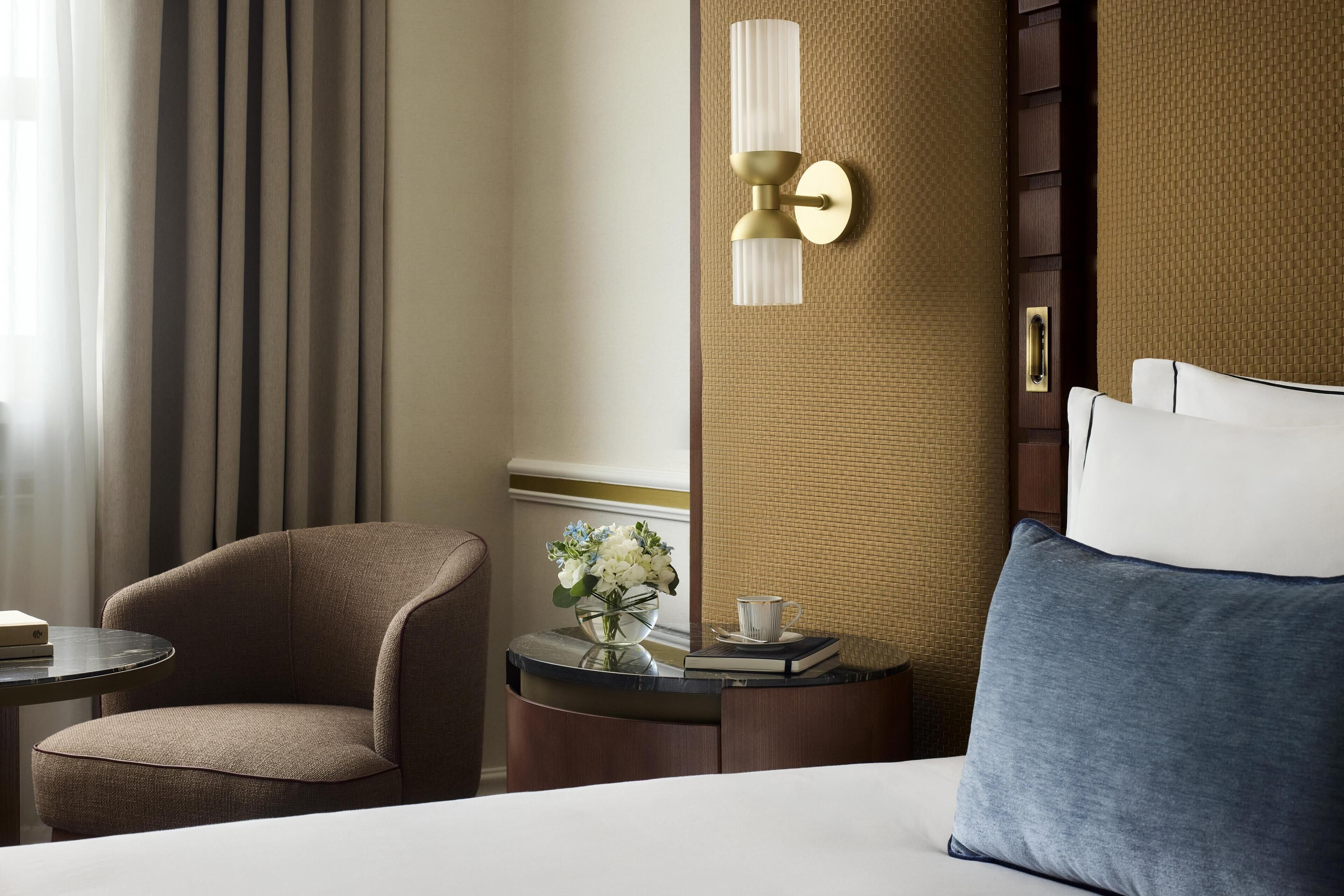 A hotel room with a bed, a chair, a small table with a vase of flowers, a cup, and a book.