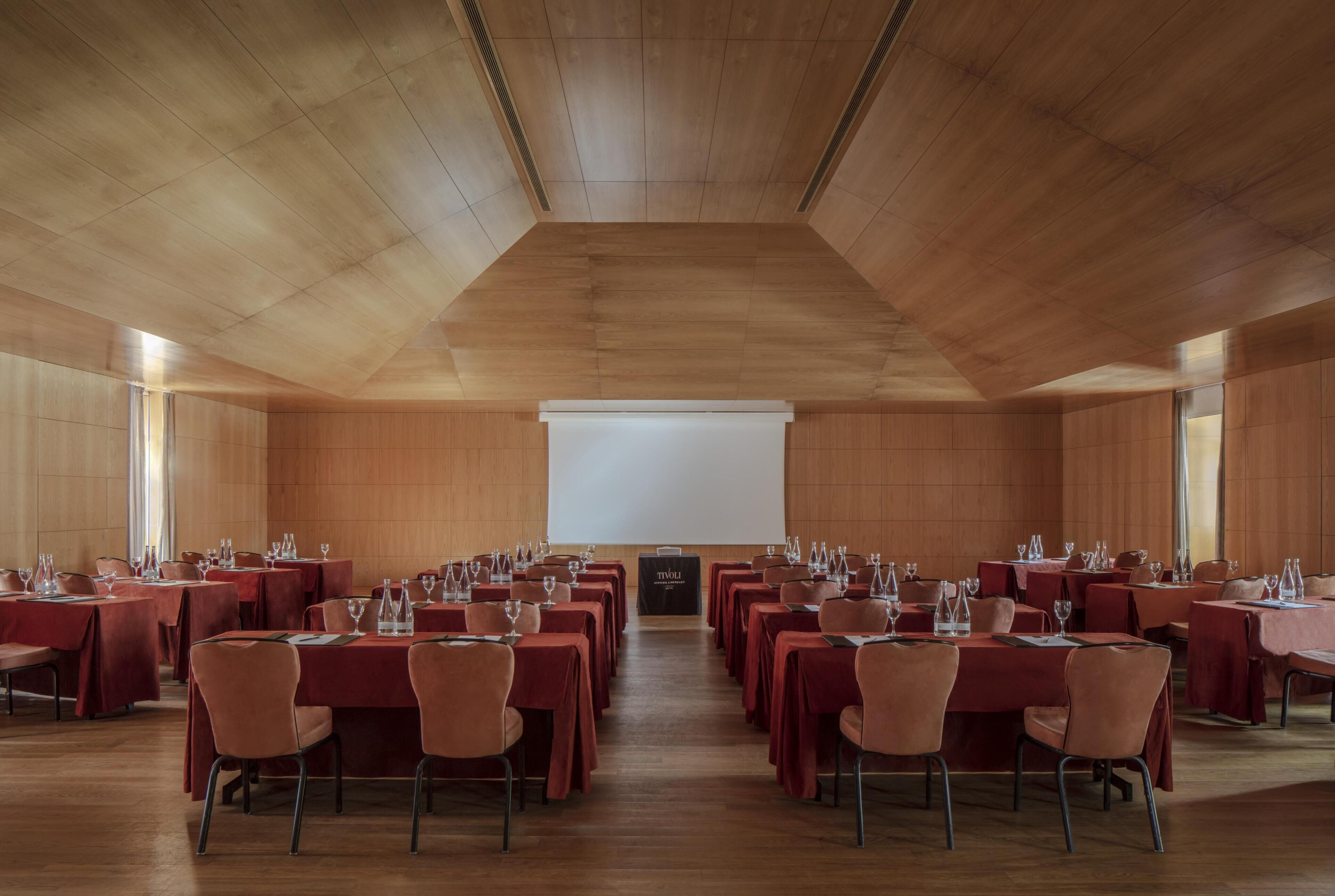 Un salón de conferencias con mesas y sillas dispuestas en filas, una pantalla de proyector y techo de madera.