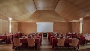 Un salón de conferencias con mesas y sillas dispuestas en filas, una pantalla de proyector y techo de madera.
