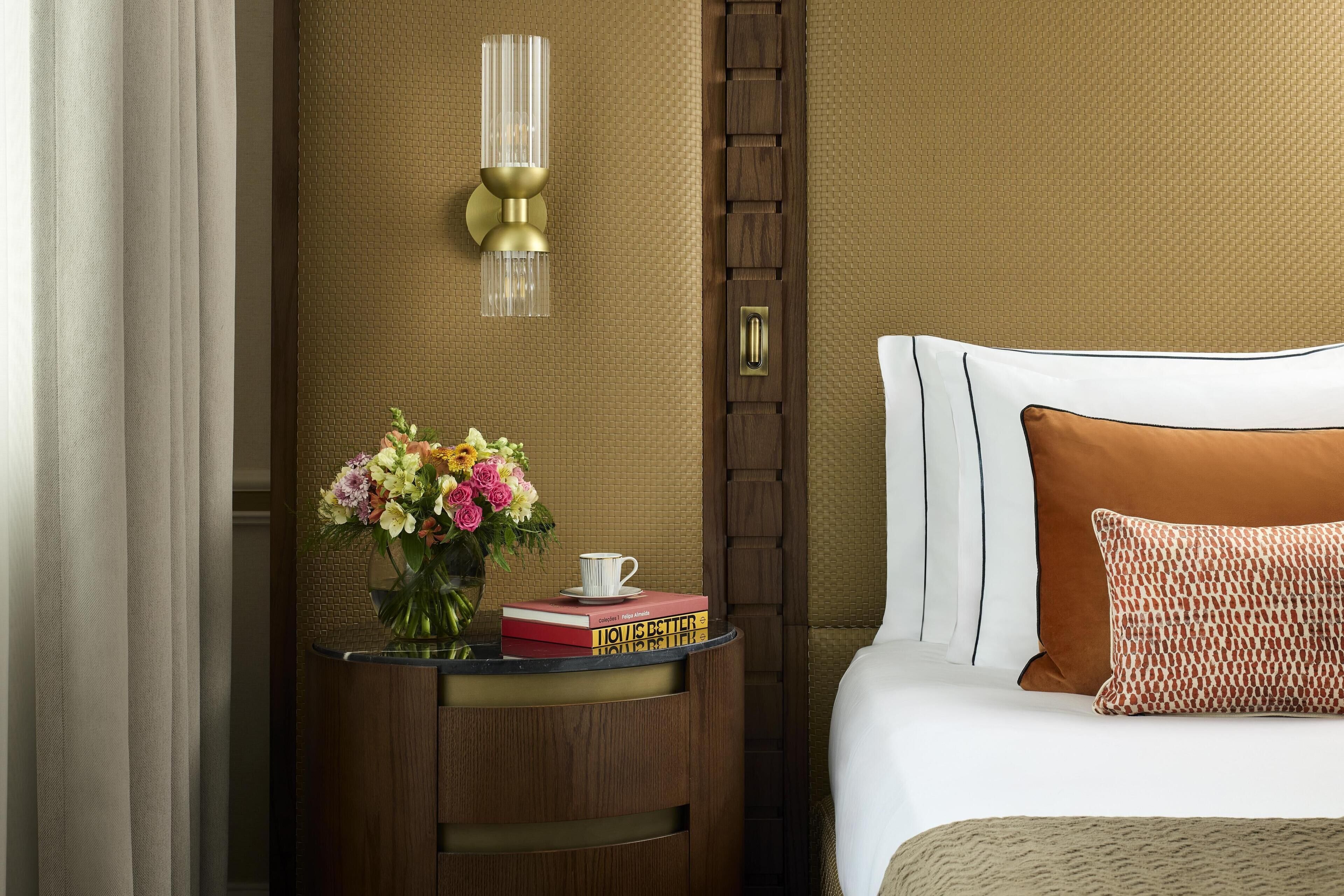 A hotel room with a wooden sideboard, a vase of flowers, a stack of books, a lamp, and a bed with white and brown bedding.