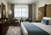 A hotel room with a large bed, a desk with a lamp, two chairs, a small table with flowers, and a large window with curtains.