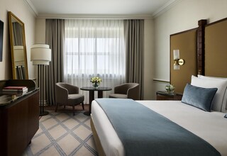A hotel room with a large bed, a desk with a lamp, two chairs, a small table with flowers, and a large window with curtains.