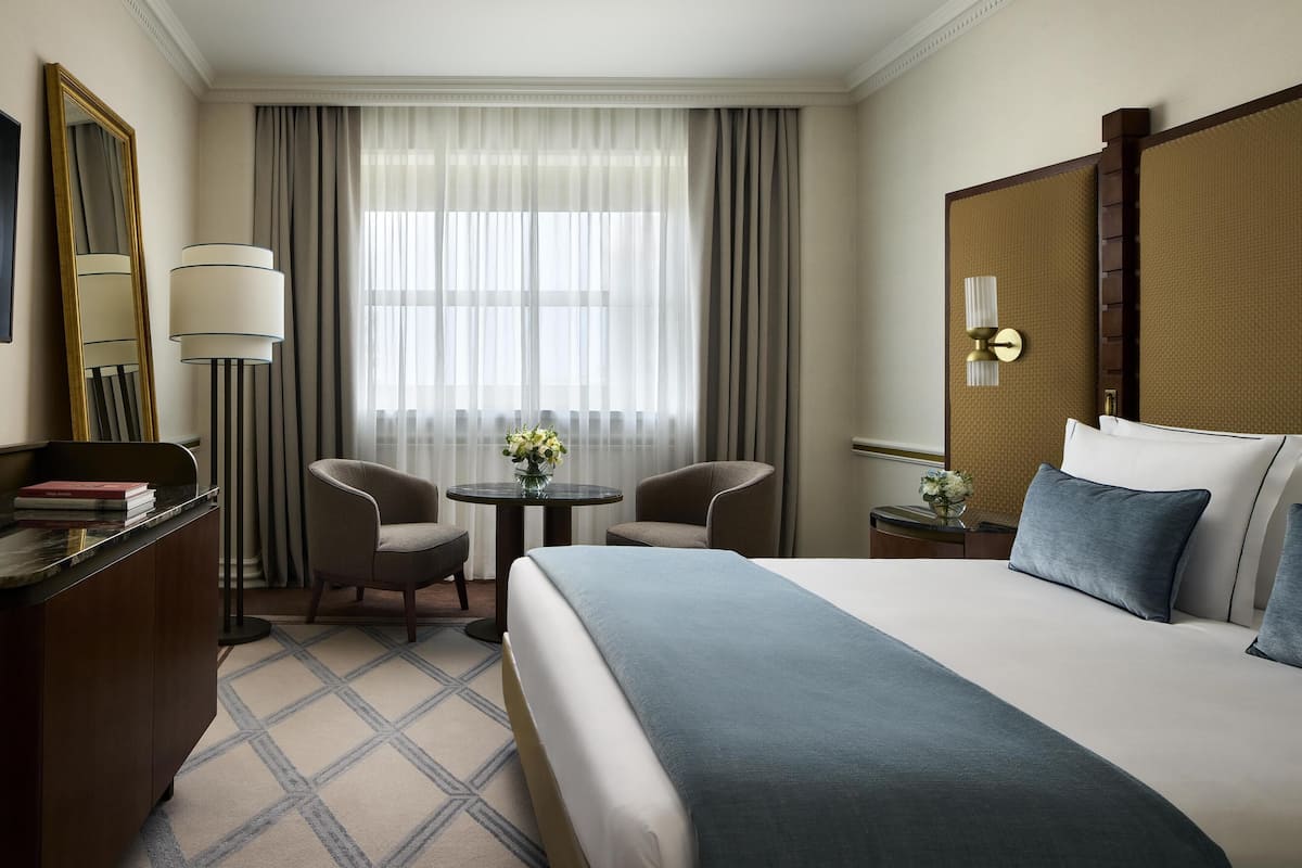 Ein Hotelzimmer mit einem großen Bett, einem Schreibtisch mit Lampe, zwei Stühlen, einem kleinen Tisch mit Blumen und einem großen Fenster mit Vorhängen.