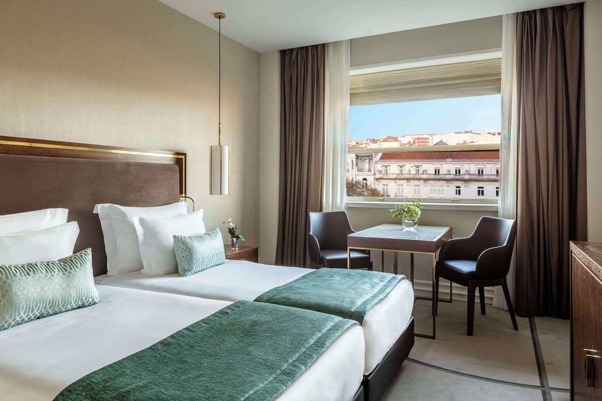 A hotel room with two beds, a small table, two chairs, a desk, and a window with a city view.
