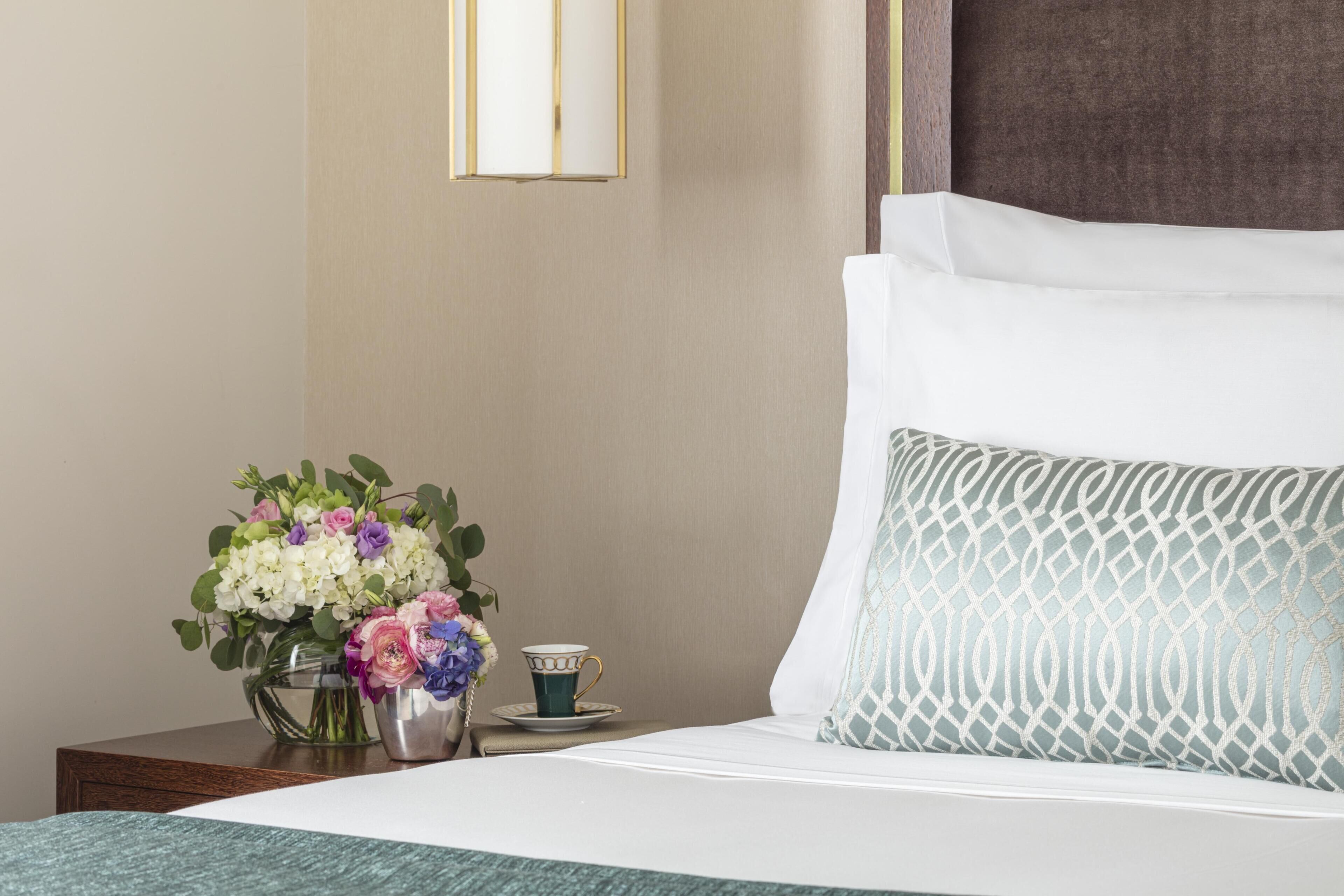 A neatly made bed with a patterned pillow, a vase with flowers, and a cup on a bedside table.
