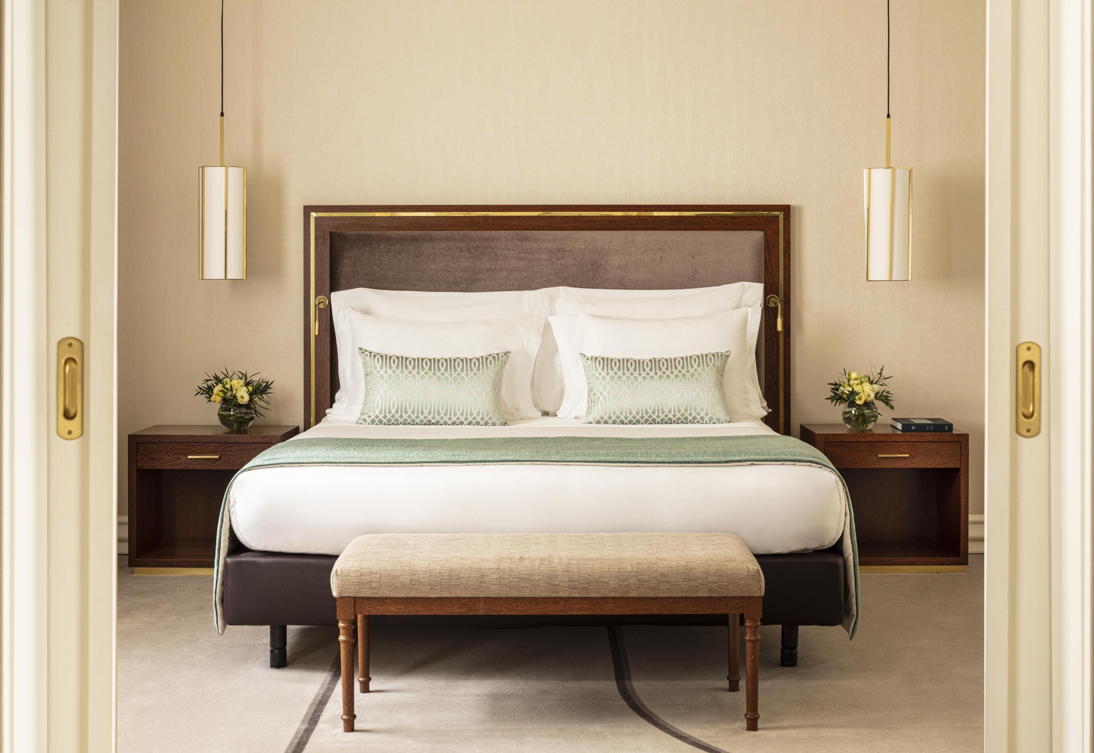 Un dormitorio con una cama grande, dos mesitas de noche, un banco y dos lámparas de pared.
