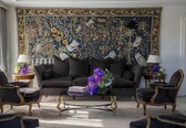 A living room with a large tapestry, a sofa, two armchairs, a coffee table, and two lamps.