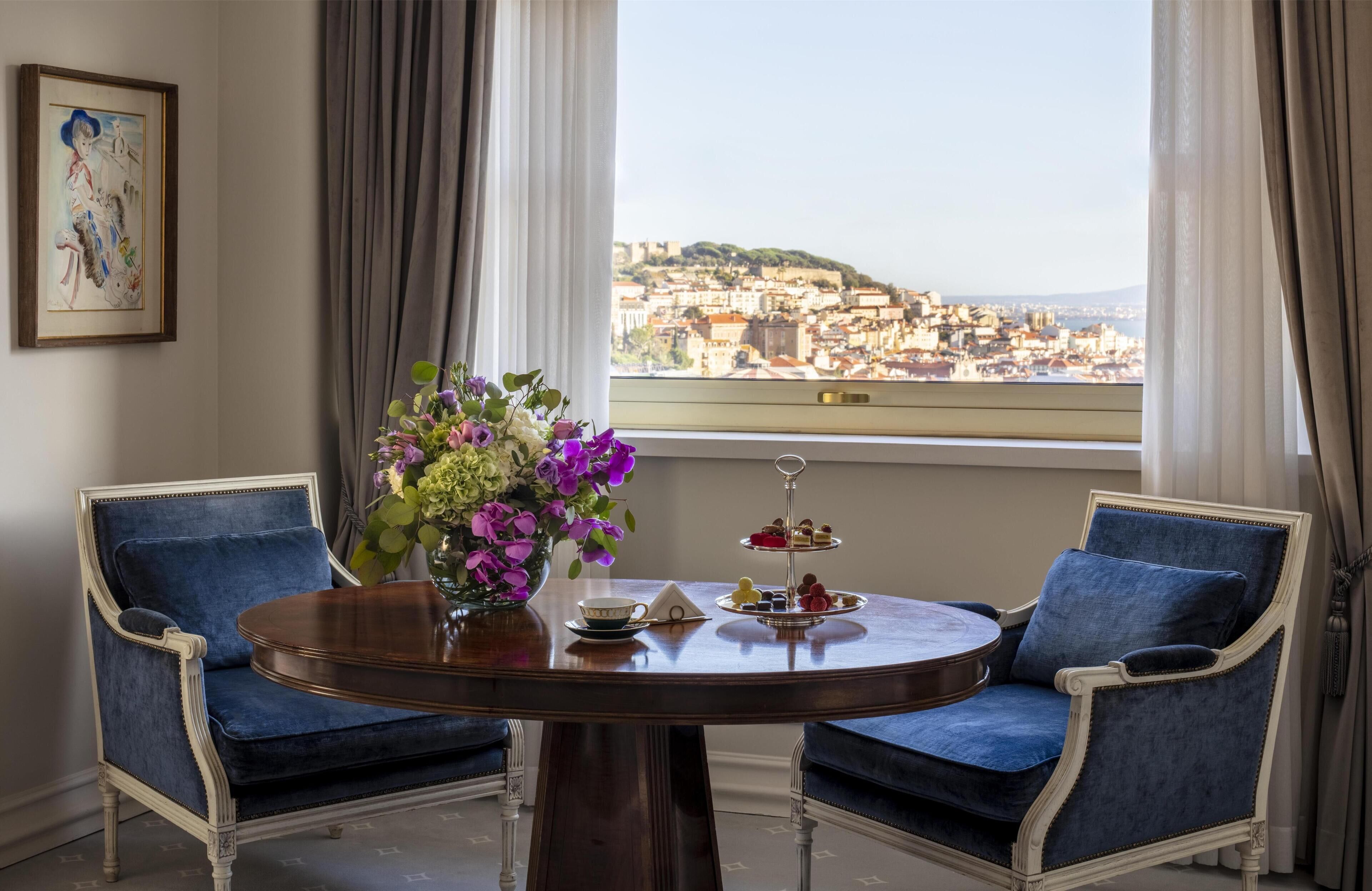 A room with a round table, two blue upholstered chairs, a vase with flowers, a tray of desserts, and a city view through a window.