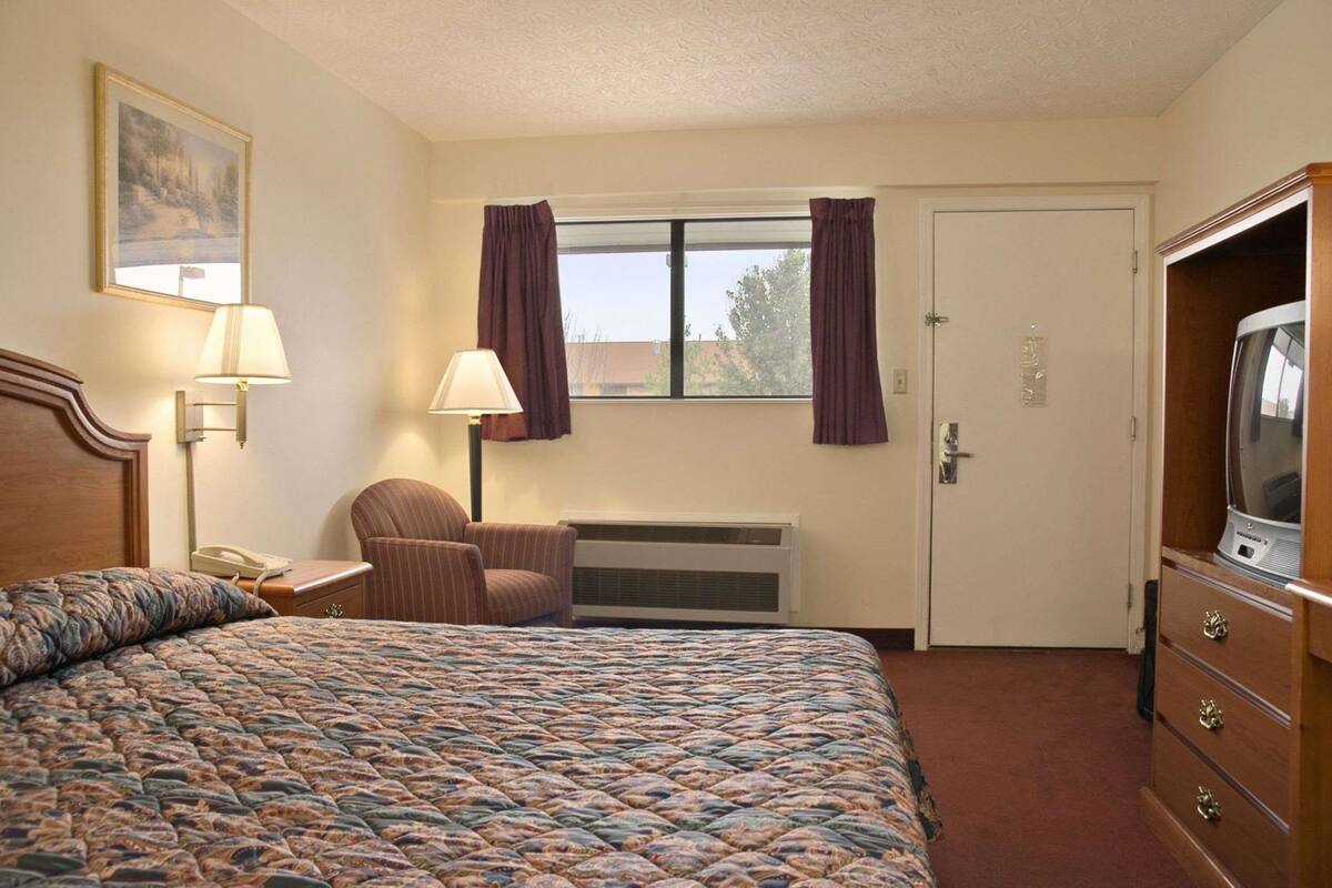 A hotel room with a bed, a chair, a desk with a lamp, a television, and a window with curtains.