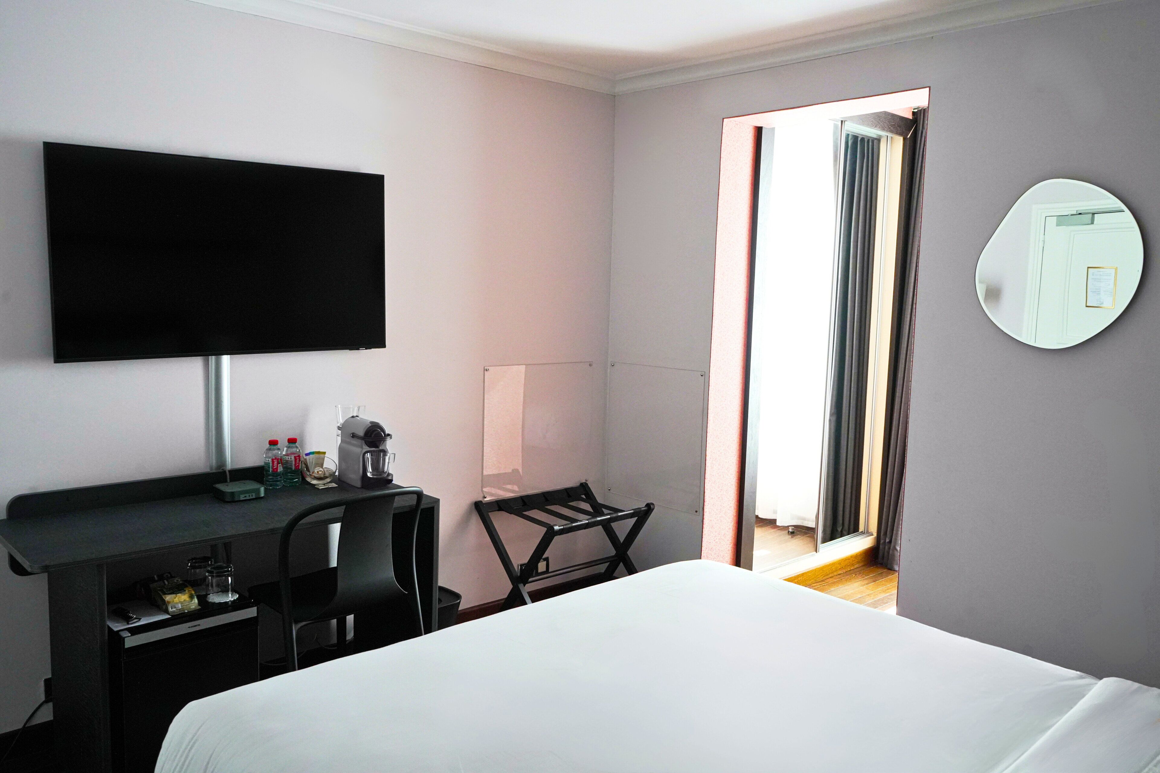 A hotel room with a bed, a desk with a flat-screen TV, a chair, a small table with a lamp, and a mirror on the wall.