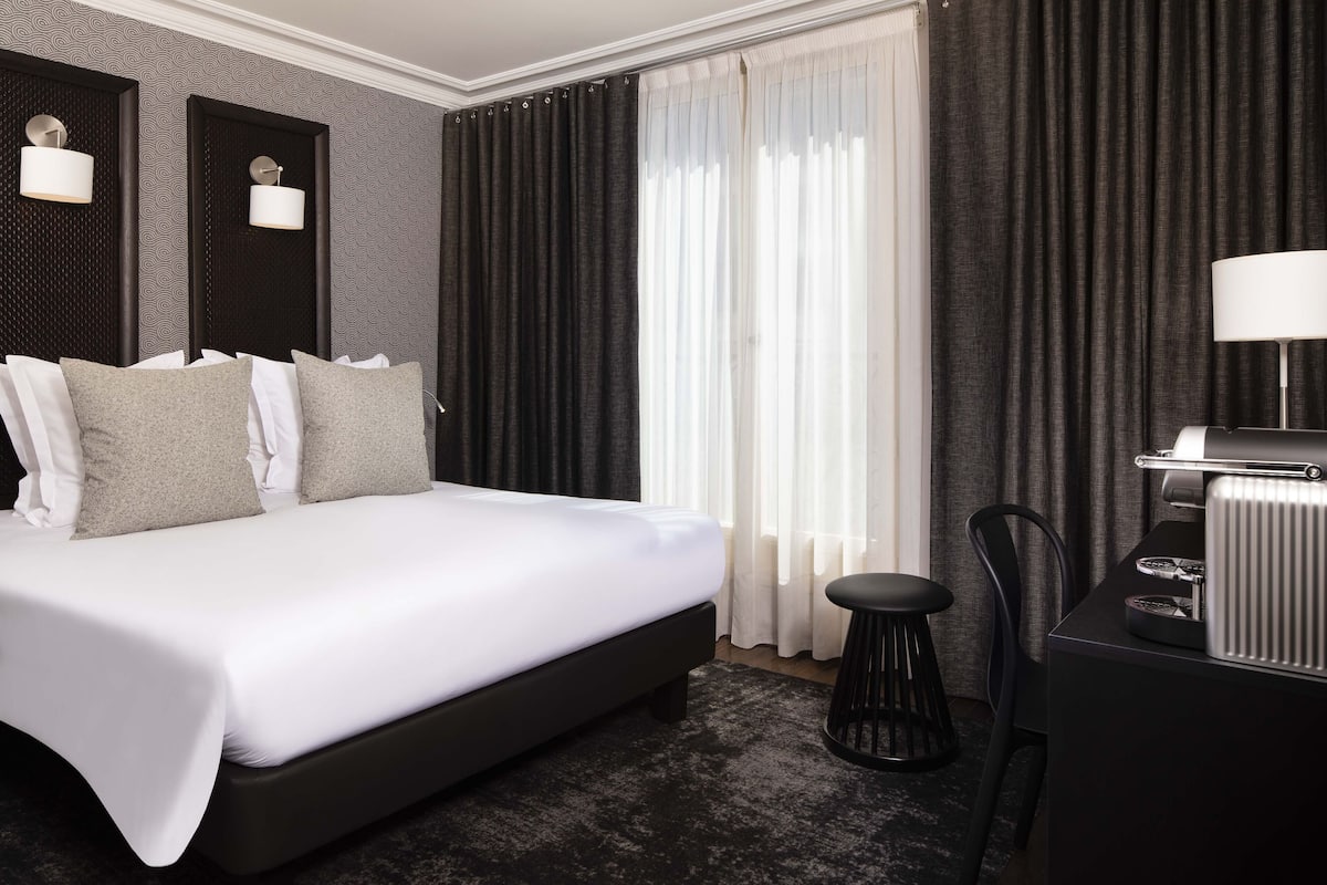 A modern hotel room with a large bed, a desk with a lamp, and a view of the outside through sheer curtains.