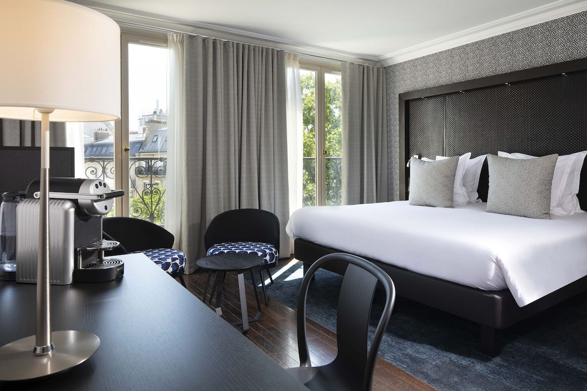 A hotel room with a bed, a desk with a coffee machine, a chair, and a view of the outdoors.
