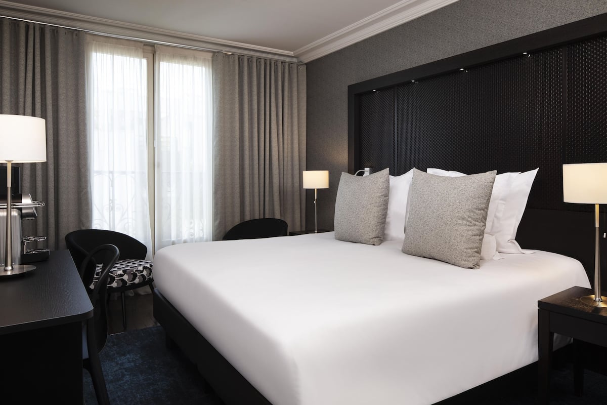 A hotel room with a large bed, two bedside tables, a desk, a chair, and a window with curtains.